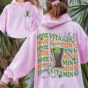 Puede incluir: Sudadera con capucha rosa claro con un dise&ntilde;o gr&aacute;fico en la parte delantera y trasera. La parte delantera muestra un vaso de cerveza con un tr&eacute;bol y el texto "100% Vitamin G". La parte trasera presenta un vaso de cerveza, una herradura y el texto "100% Vitamin G".