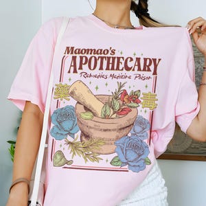 May include: A light pink t-shirt featuring a graphic design for "Maomao's Apothecary." The design includes a mortar and pestle, blue roses, and text in English and Chinese. The shirt is a casual, oversized fit.