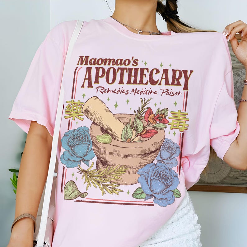 May include: A light pink t-shirt featuring a graphic design for "Maomao's Apothecary." The design includes a mortar and pestle, blue roses, and text in English and Chinese. The shirt is a casual, oversized fit.