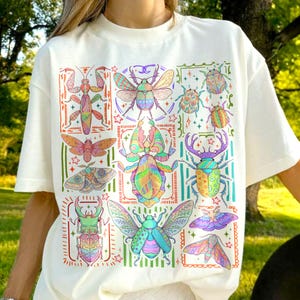 May include: A cream-colored short-sleeve t-shirt featuring a large, colorful graphic print of various insects, including beetles, moths, and a praying mantis. The insects are decorated with intricate, vibrant patterns in shades of pink, blue, green, and yellow.