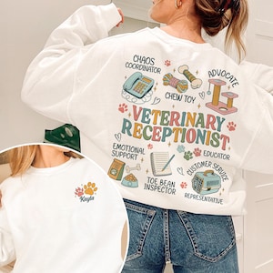 May include: A white long-sleeve sweatshirt featuring a back graphic with the text VETERINARY RECEPTIONIST, CHAOS COORDINATOR, ADVOCATE, EMOTIONAL SUPPORT, EDUCATOR, CUSTOMER SERVICE REPRESENTATIVE, and TOE BEAN INSPECTOR, alongside illustrations of a phone, chew toy, cat tree, notepad, food bag, and pet carrier. The front features a small paw print graphic with the name Kayla.