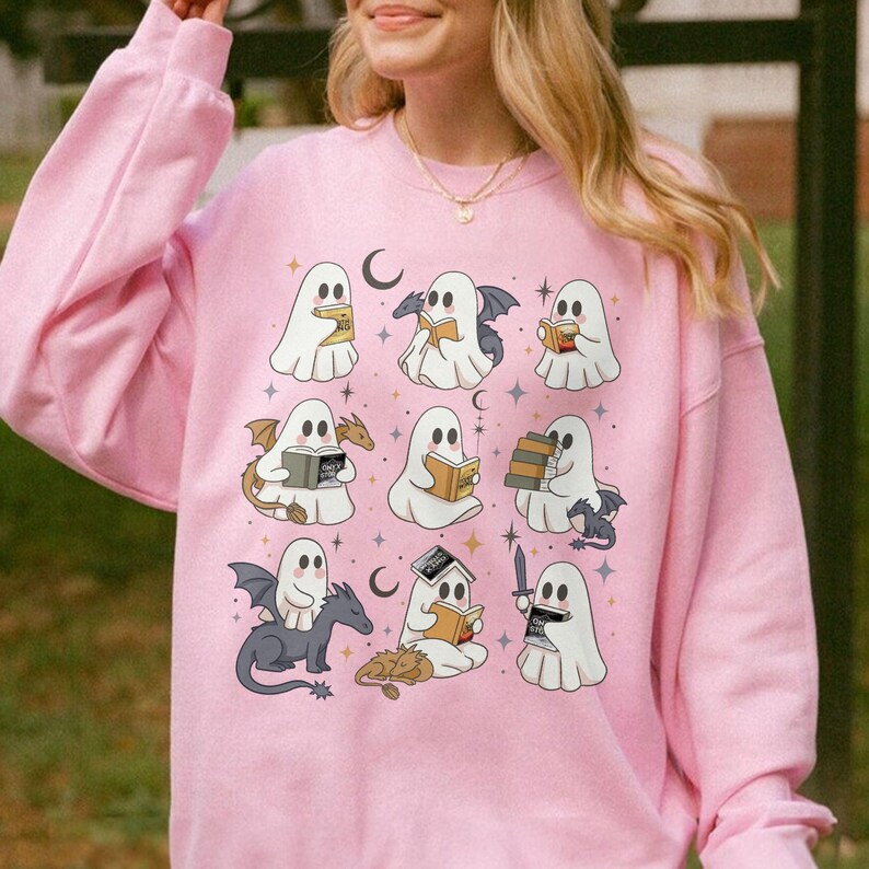 May include: Pale pink sweatshirt with a repeating pattern of cartoon ghosts reading books, some with dragon wings or riding dragons. The design includes moons, stars, and a sword. A cosy and whimsical design for Halloween or book lovers.