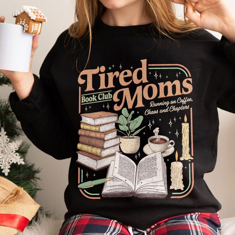 Puede incluir: Sudadera negra con un dise&ntilde;o de "Tired Moms Book Club" con ilustraciones de libros, una planta, caf&eacute; y velas. Tambi&eacute;n incluye el texto "Running on Coffee, Chaos and Chapters".