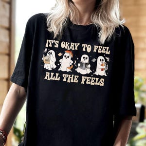 以下が含まれることがあります： 白い幽霊が本を読んでいる白いグラフィックがプリントされた黒いTシャツ。幽霊の下に「It's okay to feel all the feels」というテキストが白でプリントされています。