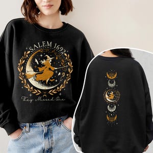 May include: Black sweatshirt featuring a graphic of a witch flying on a broomstick in front of a crescent moon. The graphic includes the text "Salem 1692" and "They Missed One". The back of the sweatshirt has a graphic of three moons with a witch flying on a broomstick in front of the middle moon.