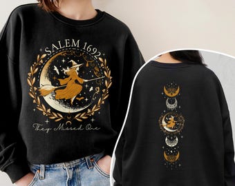 May include: A black long-sleeve sweatshirt featuring a graphic of a witch on a broomstick in front of a crescent moon, surrounded by a gold leaf wreath. The text reads SALEM 1692 and They Missed One. The back displays a vertical moon phase design.