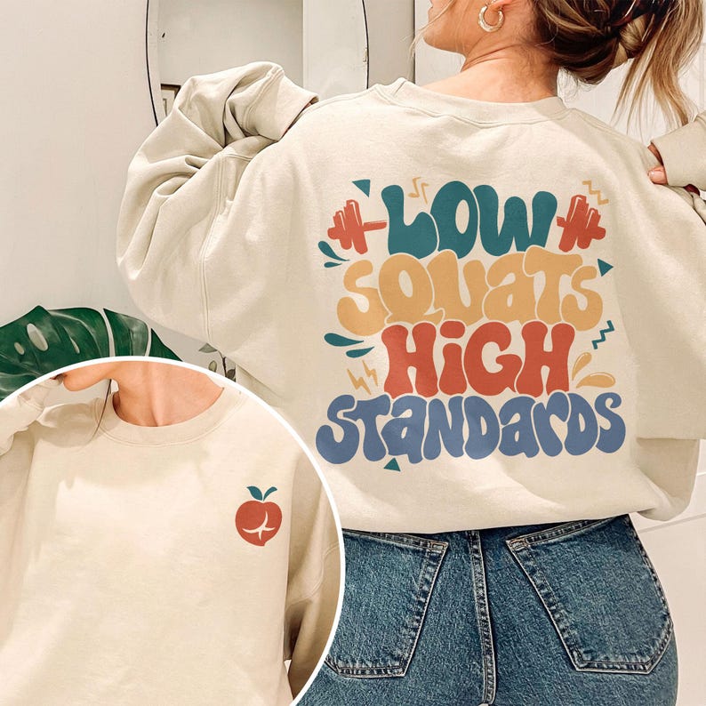 May include: A cream colored sweatshirt with the text "Low Squats High Standards" in colorful, retro-style lettering. The sweatshirt has a small apple graphic on the left sleeve.