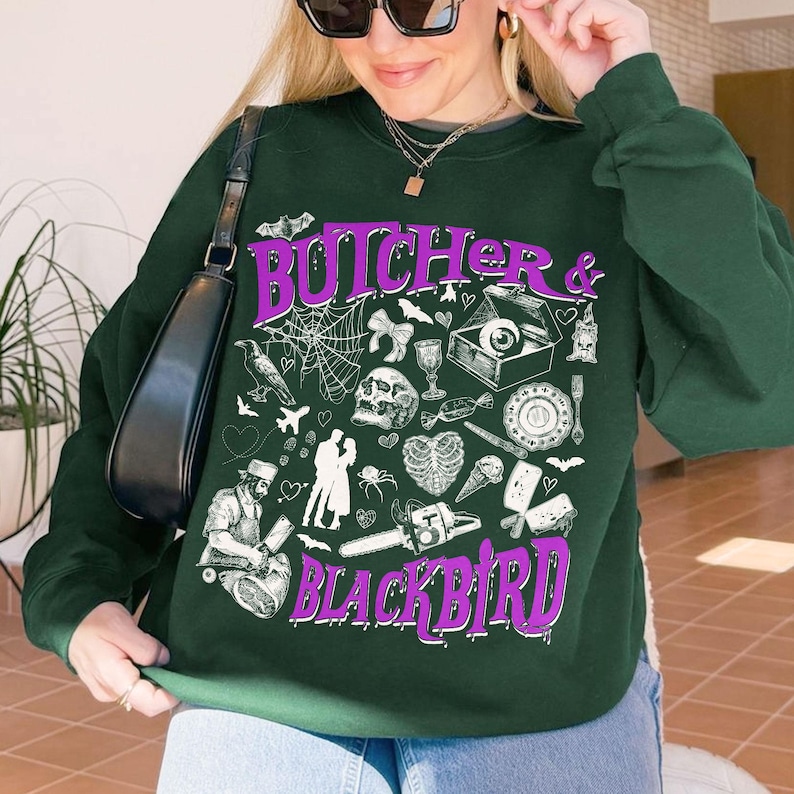 May include: Dark green sweatshirt with the text "BUTCHER & BLACKBIRD" in purple, along with various spooky illustrations. The design includes a skull, spiderwebs, and other gothic-themed elements. The sweatshirt is a crew neck style.