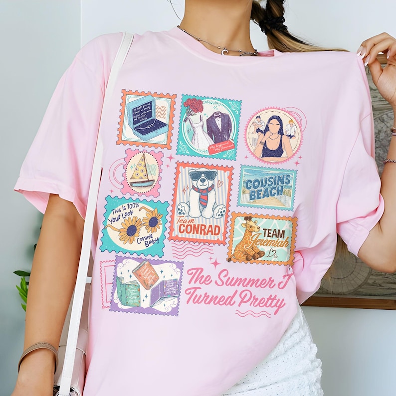 May include: A light pink t-shirt featuring a colorful stamp design. The stamps depict various illustrations, including a laptop, wedding attire, a sailboat, a bear wearing sunglasses, and the text "The Summer I Turned Pretty."
