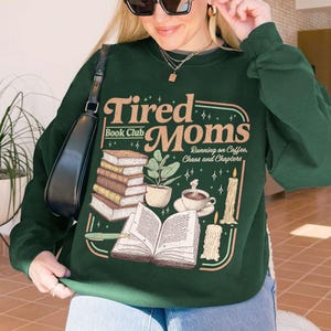 Puede incluir: Sudadera verde oscuro con el texto "Tired Moms Book Club" en un dise&ntilde;o retro. El gr&aacute;fico incluye ilustraciones de libros, una planta, una taza de caf&eacute; y velas. La sudadera es de cuello redondo.