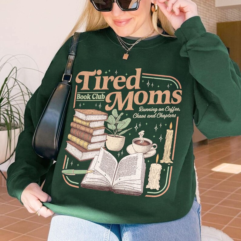 Puede incluir: Sudadera verde oscuro con el texto "Tired Moms Book Club" en un dise&ntilde;o retro. El gr&aacute;fico incluye ilustraciones de libros, una planta, una taza de caf&eacute; y velas. La sudadera es de cuello redondo.