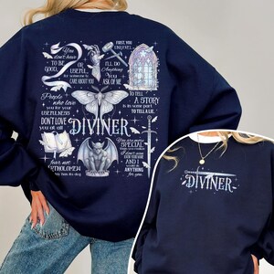 May include: A navy blue sweatshirt featuring a large back graphic with a moth, a gargoyle, a sword, a gothic window, and text including "DIVINER" and "You don't have to be good, or useful, for someone to care about you." The front has a smaller "DIVINER" logo.