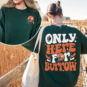 以下が含まれることがあります： オレンジと白で「ONLY HERE FOR BURROW」と書かれた緑色のスウェットシャツ。フットボールと、9番のフットボールヘルメットが描かれている。