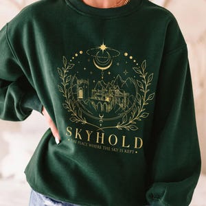 May include: A dark green sweatshirt with a gold graphic design of a castle, stars, and a crescent moon. The text "SKYHOLD" is below the graphic, with the words "THE PLACE WHERE THE SKY IS KEPT" underneath.