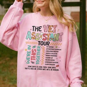May include: A light pink long-sleeve sweatshirt featuring the text THE VET ASSISTANT TOUR in colorful patterned letters. Below, the text reads WE'RE IN THIS TOGETHER with a list of tasks like PET SOOTHIN and the phrase SOME DAYS I'LL HEAL THEM.
