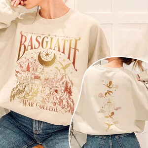 K&ouml;nnte beinhalten: Ein beiger Pullover mit einem goldenen und roten Grafikdruck eines Schlosses und Drachen. Der Text "Basgiath War College" ist auf der Vorderseite des Pullovers gedruckt. Die R&uuml;ckseite des Pullovers hat einen goldenen und roten Grafikdruck von Drachen.