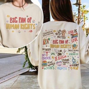 May include: A cream-colored crewneck sweatshirt featuring the text BIG FAN OF HUMAN RIGHTS on the front. The back displays a collage of colorful graphics and slogans including FEMINISM FOR EVERYONE, BLACK LIVES MATTER, and science icons.