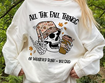 May include: A cream-colored crewneck sweatshirt featuring a graphic of a skeleton wearing a patterned bucket hat, holding a coffee cup, and making a peace sign. The text reads "ALL THE FALL THINGS" and "OR WHATEVER BLINK-182 SAID" with autumn leaves.