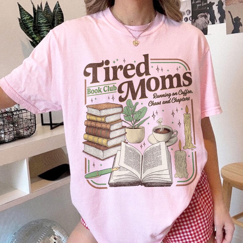 Puede incluir: Camiseta rosa claro con un dise&ntilde;o de "Tired Moms Book Club" con ilustraciones de libros, una planta, caf&eacute; y velas. Tambi&eacute;n incluye el texto "Running on Coffee, Chaos and Chapters".