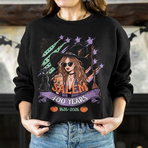 May include: A black long-sleeve sweatshirt featuring a graphic of a witch wearing a hat and sunglasses, holding a drink. Beside her is a black cat and a broomstick. The text reads SALEM 400 YEARS 1626-2026, accented with two orange pumpkins.