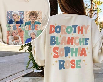 Könnte beinhalten: Cremefarbenes Sweatshirt mit einer Collage von vier Frauen und dem Text "STAY GOLDEN" auf der Vorderseite. Auf der Rückseite des Sweatshirts stehen die Namen "DOROTHY BLANCHE SOPHIA ROSE" in bunten Blockbuchstaben.