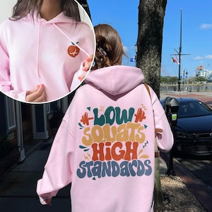 May include: Pink hoodie with a graphic on the back that says "Low Squats High Standards" in colorful, retro-style lettering.