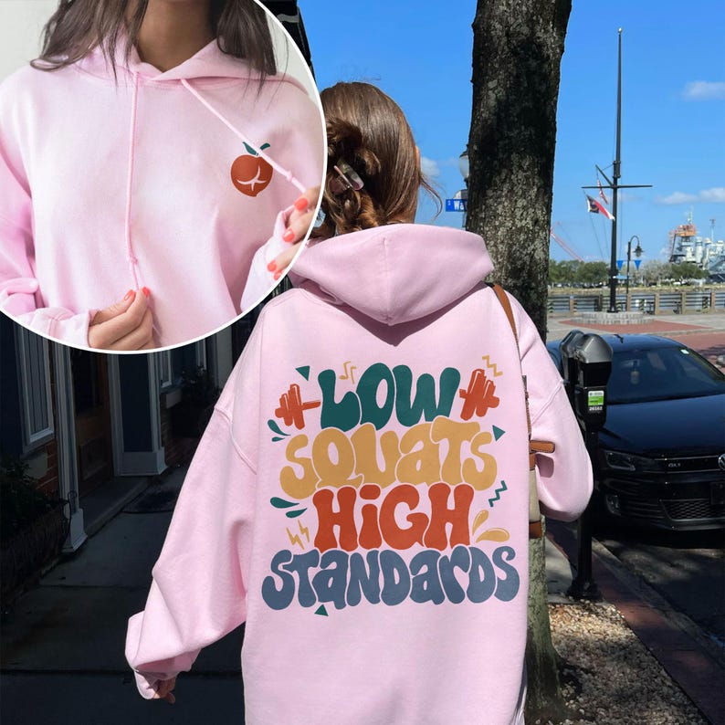 May include: Pink hoodie with a graphic on the back that says "Low Squats High Standards" in colorful, retro-style lettering.