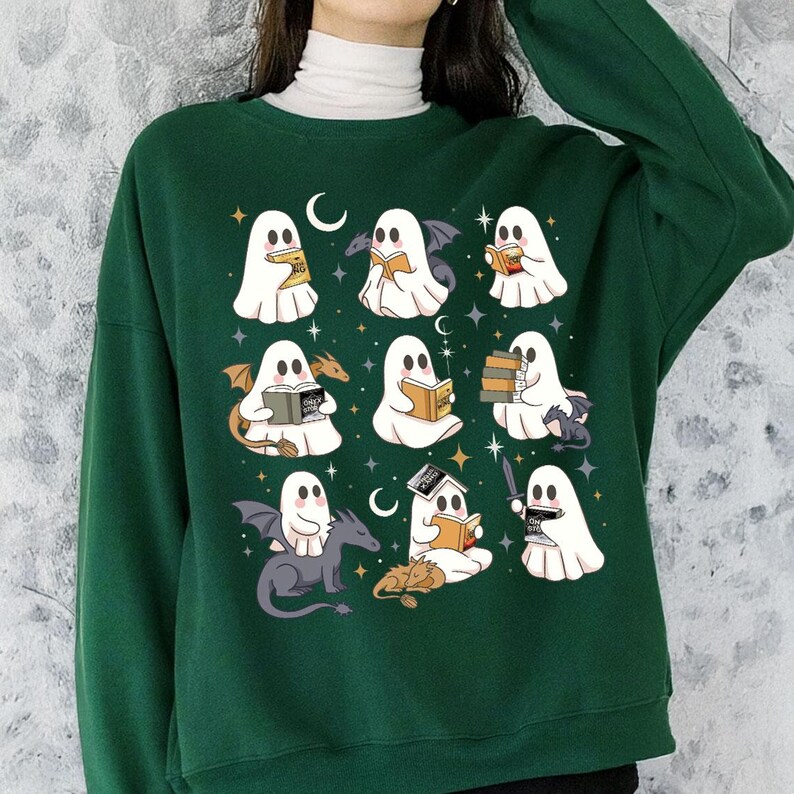 May include: Dark green sweatshirt with a repeating pattern of cartoon ghosts reading books, with bats, dragons, and moons. The ghosts are white with black eyes and are holding books of various colours. The design is repeated across the front of the sweatshirt.