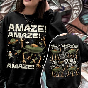 May include: Black sweatshirt with a whimsical space-themed design. The front features the word "AMAZE!" repeated, along with illustrations of a planet, rocket, astronaut, and a robot-like alien. The back has alien illustrations and phrases like "JAZZ HANDS" and "BE CAREFUL YOU ARE FRIEND NOW."