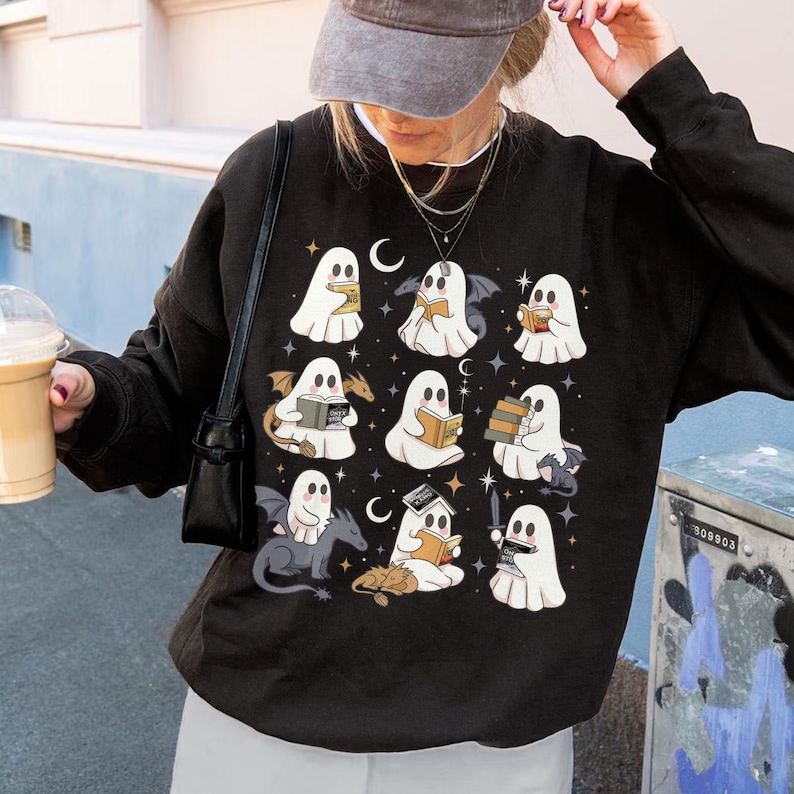 May include: Black sweatshirt with a pattern of white ghost illustrations reading books, with bats, dragons, and crescent moons. The design includes stars and small details, creating a whimsical and spooky aesthetic.