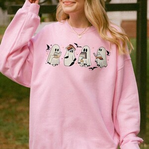 Puede incluir: Una sudadera rosa con un gr&aacute;fico de cuatro fantasmas leyendo libros. Los fantasmas son blancos y tienen ojos negros. El texto en la sudadera es "Boo-kworms".