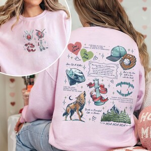 K&ouml;nnte beinhalten: Rosafarbener Pullover mit einem grafischen Druck aus verschiedenen Bildern und Texten. Die Bilder beinhalten einen Diamanten, einen Wolf, eine Schachfigur, einen Baseballhandschuh, einen Baseball und eine Bergkette. Der Text beinhaltet "Team Edward", "Team Jacob", "Sin of a Killer", "Take the night without the dark, we'd never see the stars", "Death is Peaceful, Life is Harder", "And so the lion fell in love with the lamb", "Twilight, main another ending, no more than perfect, no more than it allows has to end", and "HOA HOA HOA".