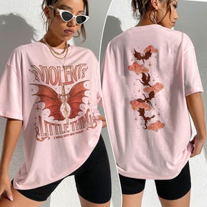 May include: Pink oversized t-shirt with a graphic print of a red dragon with a sword and the text "VIOLENT LITTLE THING I WILL NOT SIN TODAY". The back of the shirt has a graphic print of five black dragons flying in the clouds.