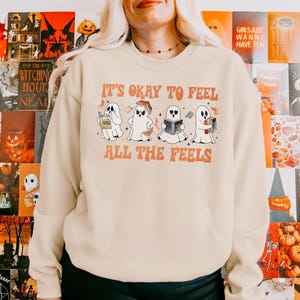 以下が含まれることがあります： クリーム色のスウェットシャツに、「It's okay to feel all the feels」という文字が前面にプリントされています。文字はオレンジと茶色です。白い体に黒い目と口をした漫画の幽霊が4体、本を読んでいます。