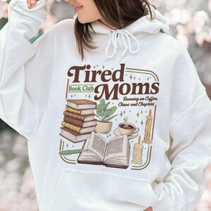 Puede incluir: Sudadera con capucha blanca con un gr&aacute;fico marr&oacute;n y verde que dice "Tired Moms Book Club". El dise&ntilde;o incluye ilustraciones de libros, una planta, caf&eacute; y una vela. Tambi&eacute;n incluye el texto "Running on Coffee, Chaos and Chapters".