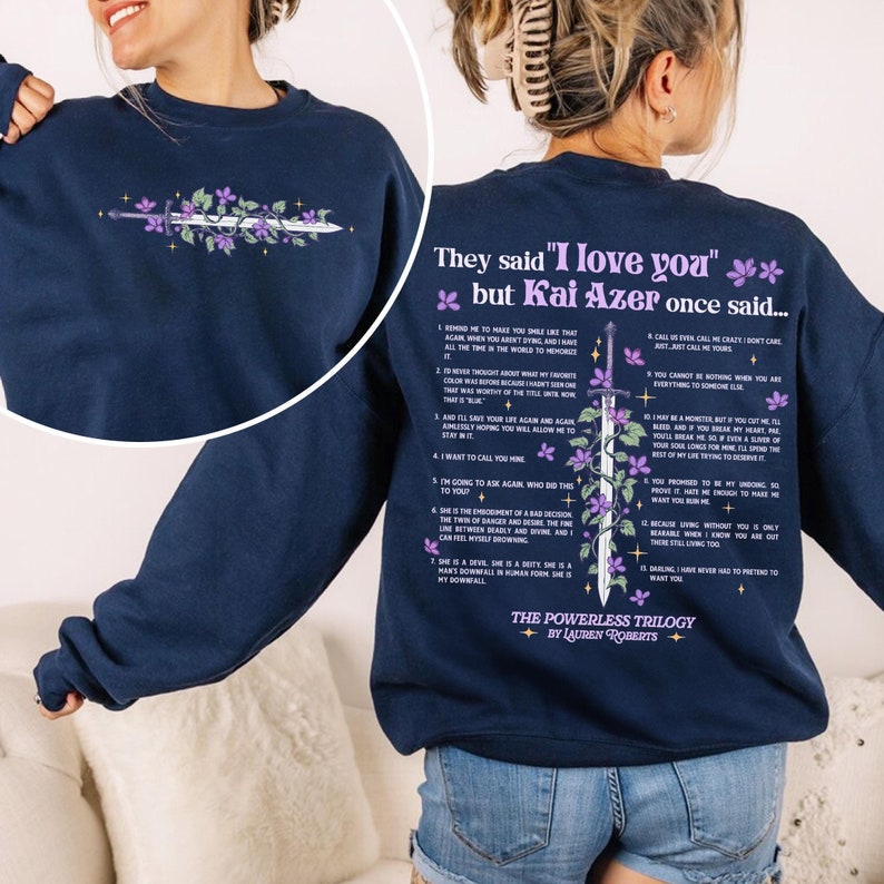 May include: A navy blue sweatshirt with a quote from the book "The Powerless Trilogy" by Lauren Roberts. The quote reads "They said "I love you" but Kai Azer once said..." and includes a list of 12 points. The sweatshirt features a purple floral design with a sword.