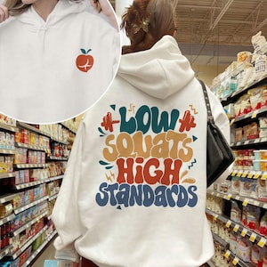 May include: White hooded sweatshirt with a graphic print on the back that says "Low Squats High Standards" in colorful, retro-style lettering. The front of the sweatshirt has a small red apple graphic.