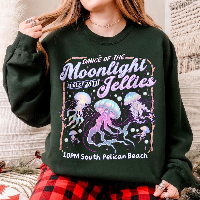 Puede incluir: Sudadera verde oscuro con un dise&ntilde;o gr&aacute;fico de medusas en tonos morados, azules y rosas. El texto dice "Dance of the Moonlight Jellies, August 28th, 10PM South Pelican Beach."