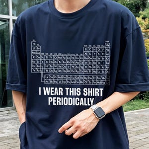 May include: Navy blue t-shirt featuring a periodic table design in white. The shirt includes the text "I WEAR THIS SHIRT PERIODICALLY". The shirt is a short-sleeved, casual style.