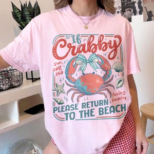 以下が含まれることがあります： ピンク色のTシャツ。カニ、ヒトデ、貝殻のグラフィックデザインが特徴です。「If Crabby, Please Return to the Beach」のテキスト。デザインには「sun, salt, sea」と「feeling beachy」の文字が含まれています。