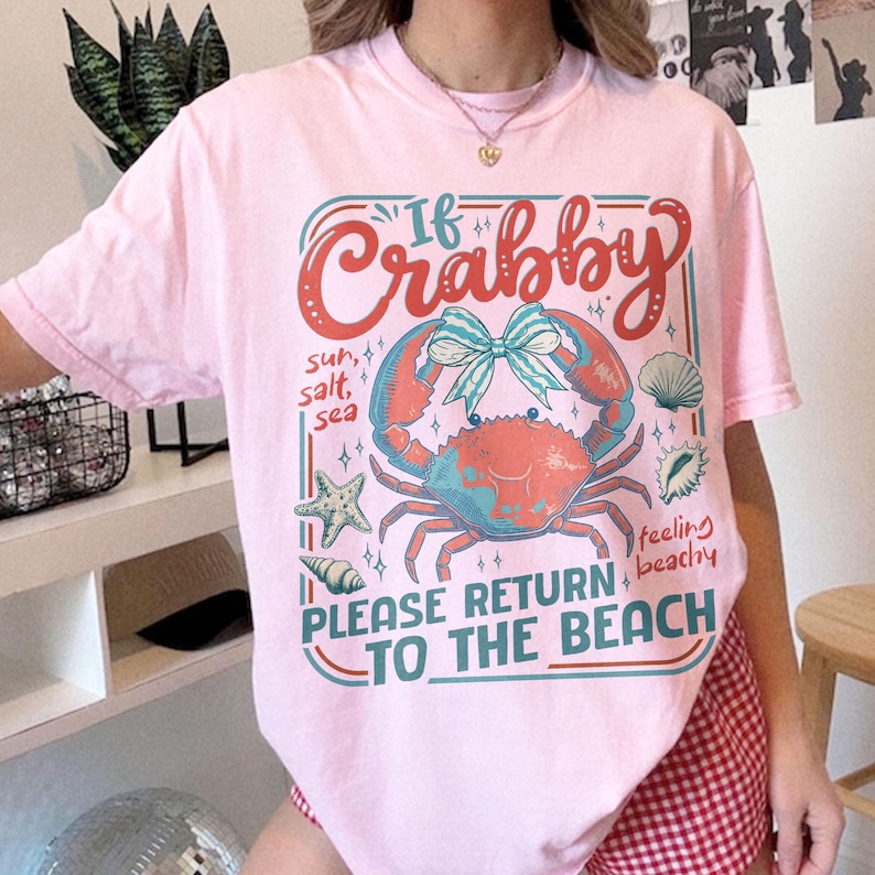 以下が含まれることがあります： ピンク色のTシャツ。カニ、ヒトデ、貝殻のグラフィックデザインが特徴です。「If Crabby, Please Return to the Beach」のテキスト。デザインには「sun, salt, sea」と「feeling beachy」の文字が含まれています。