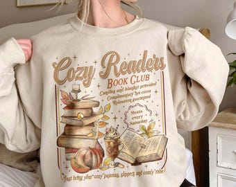 May include: A beige crewneck sweatshirt featuring a graphic print with the text Cozy Readers BOOK CLUB, a stack of books, a pumpkin, a mug of hot cocoa, and an open book, surrounded by decorative stars and autumn leaves on the front.