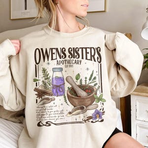 May include: A beige crewneck sweatshirt featuring a dark brown graphic print titled Owens Sister's Apothecary Est 1995. The design includes a purple liquid bottle, a wooden mortar and pestle, cinnamon sticks, herbs, and handwritten text.