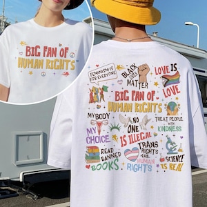 May include: A white t-shirt featuring a graphic print on the front and back. The front displays the text BIG FAN OF HUMAN RIGHTS. The back features various colorful illustrations and text including FEMINISM IS FOR EVERYONE, BLACK LIVES MATTER, LOVE IS LOVE, MY BODY MY CHOICE, NO ONE IS ILLEGAL, READ BANNED BOOKS, TRANS RIGHTS ARE HUMAN RIGHTS, and SCIENCE IS REAL.