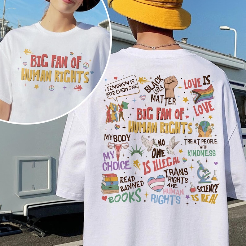 May include: A white t-shirt featuring a graphic print on the front and back. The front displays the text BIG FAN OF HUMAN RIGHTS. The back features various colorful illustrations and text including FEMINISM IS FOR EVERYONE, BLACK LIVES MATTER, LOVE IS LOVE, MY BODY MY CHOICE, NO ONE IS ILLEGAL, READ BANNED BOOKS, TRANS RIGHTS ARE HUMAN RIGHTS, and SCIENCE IS REAL.