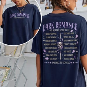 May include: Navy blue t-shirt with the text "DARK ROMANCE READER SOCIETY" in purple. The back of the shirt lists trigger warnings and other phrases in purple. A skull graphic is also visible.