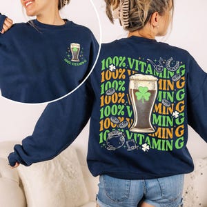 Puede incluir: Sudadera azul marino con un dise&ntilde;o de vaso de cerveza y el texto "100% Vitamin G" en la parte delantera. La parte trasera presenta un dise&ntilde;o m&aacute;s grande con un vaso de cerveza, tr&eacute;bol y texto en verde y naranja.