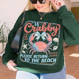 以下が含まれることがあります： カニのグラフィックと「If Crabby, Please Return to the Beach」の文字が入った濃い緑色のスウェットシャツ。デザインにはカニ、貝殻、「sun, salt, sea」と「feeling beachy」の文字が含まれています。