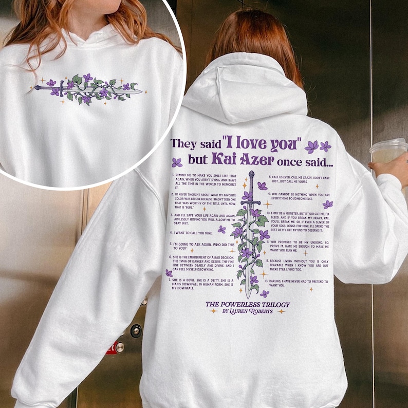 May include: White hoodie with a purple sword and flowers design. The text "They said "I love you" but Kai Azer once said..." is printed on the back of the hoodie. The text includes a list of numbered quotes. The text "The Powerless Trilogy by Lauren Roberts" is printed at the bottom of the hoodie.