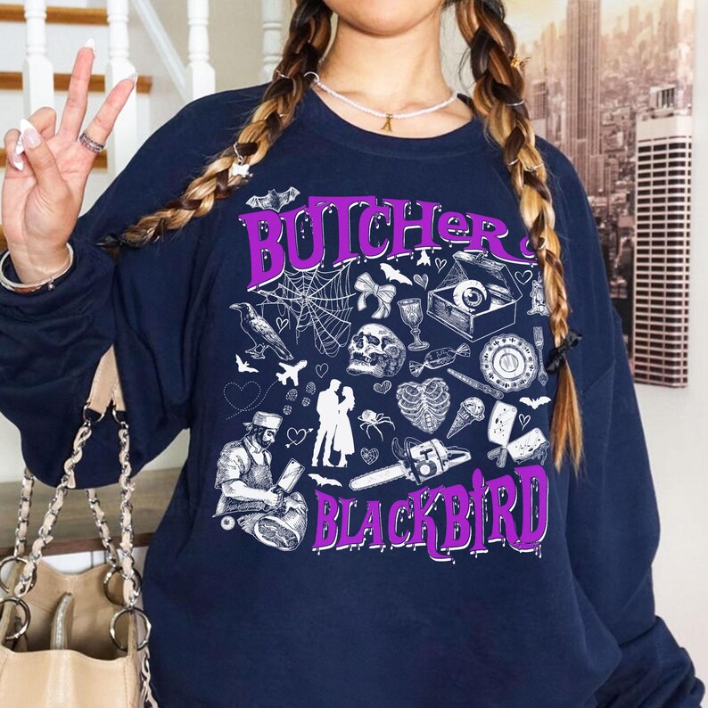 May include: Navy blue sweatshirt with a purple graphic that reads "Butcher Blackbird." The design includes illustrations of a spiderweb, skull, hearts, and a butcher. A chainsaw is also featured in the design.